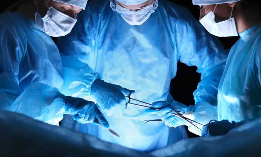 Three surgeons in blue scrubs perform fistula surgery on a patient in a sterile operating room.