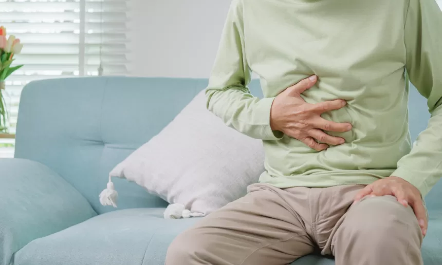 A man sits on a couch, grimacing in pain, indicating discomfort likely due to a hernia condition requiring surgery.