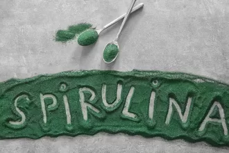 A vibrant close-up of Spirulina powder and tablets placed in a wooden bowl with a glass of green Spirulina smoothie on a rustic wooden table.