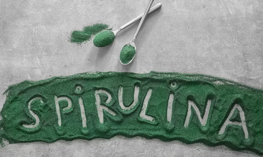 A vibrant close-up of Spirulina powder and tablets placed in a wooden bowl with a glass of green Spirulina smoothie on a rustic wooden table.